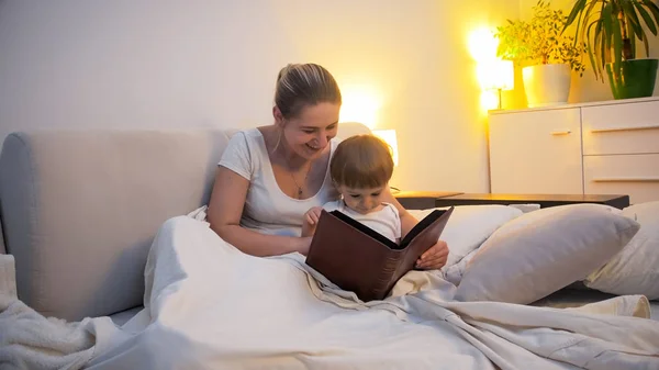 Ευτυχισμένη γυναίκα διαβάζοντας μεγάλο βιβλίο για τον γιο του νήπιο στο κρεβάτι τη νύχτα — Φωτογραφία Αρχείου