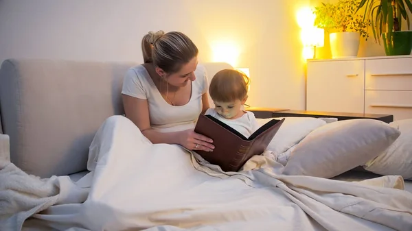 Glückliche junge Frau liest ihrem kleinen Sohn nachts im Bett eine Geschichte vor — Stockfoto