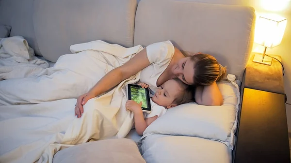 Joven madre con su hijo pequeño acostado en la cama con una tableta digital — Foto de Stock