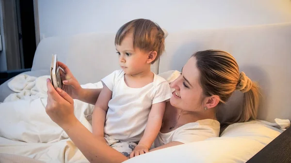 Retrato de la joven madre niño pequeño hijo sosteniendo tableta digital en la cama por la noche —  Fotos de Stock