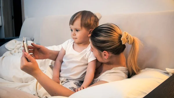 Retrato tonificado para niño lindo con madre acostada en la cama con tableta digital —  Fotos de Stock