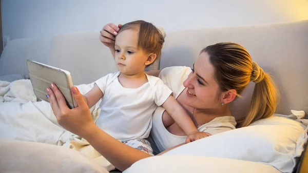 Retrato de madre sonriente con niño pequeño sosteniendo la tableta digital en la cama —  Fotos de Stock