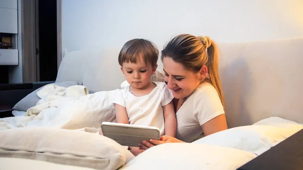 Lindo niño sentado en la cama con su madre y viendo dibujos animados en la tableta —  Fotos de Stock