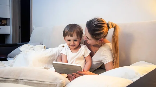 Feliz madre sonriente y niño mintiendo en la cama y viendo una película en una tableta digital —  Fotos de Stock