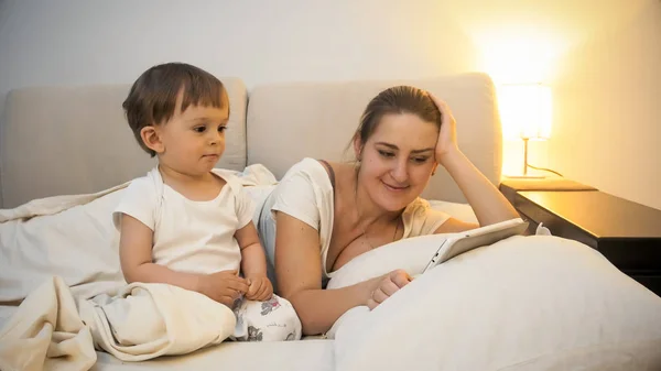 Hermosa madre joven con su hijo de 2 años de edad, acostado en la cama con la tableta digital —  Fotos de Stock