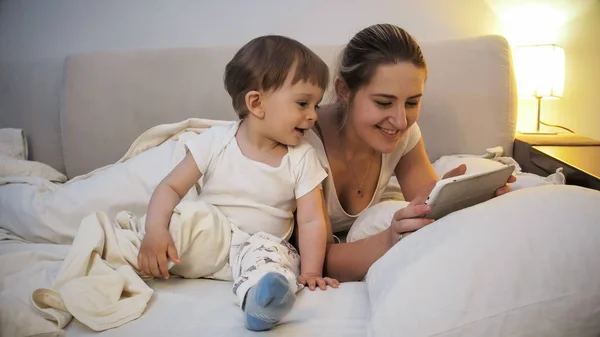 Szczęśliwy młody syn matka i dziecko w piżamie przy użyciu komputera typu tablet cyfrowy przed pójściem spać — Zdjęcie stockowe
