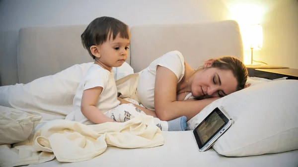 Молодая уставшая мать заснула, пока ее маленький сын смотрел мультики на цифровом планшете — стоковое фото