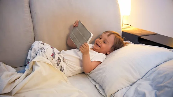 Portret szczęśliwy roześmiany toddler chłopiec leżąc w łóżku z cyfrowego tabletu — Zdjęcie stockowe