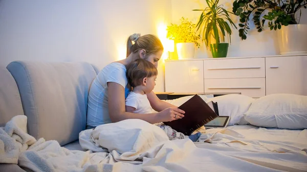 Geceleri yatakta büyük kitap okuma yürümeye başlayan çocuk oğlu ile genç anne — Stok fotoğraf