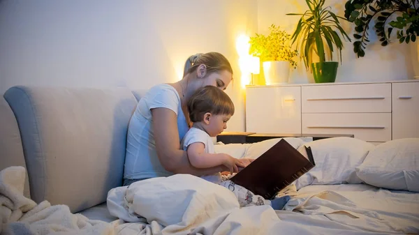 แม่สาวในชุดนอนอ่านหนังสือเล่มใหญ่ให้ลูกชายของเธอก่อนเข้านอน — ภาพถ่ายสต็อก