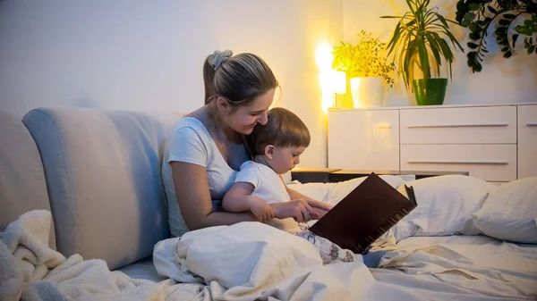 Lindo niño en pijama leyendo un gran libro con la madre en la cama —  Fotos de Stock
