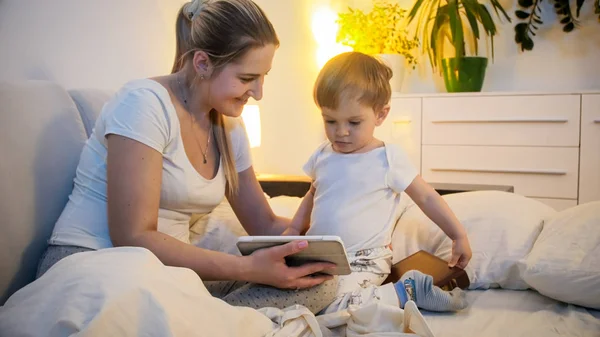 Mooie jonge vrouw in pyjama met haar 2 jaar oude zoon kijken cartoons op digitale tablet bij nacht — Stockfoto