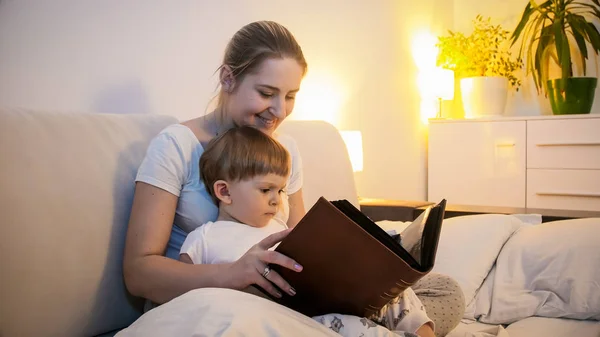 快乐的年轻母亲的画像与蹒跚学步的儿子读大书在晚上 — 图库照片