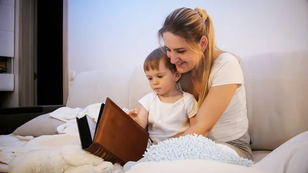 年轻的微笑的妇女的画像坐在床上与她的蹒跚学步的儿子和读大书 — 图库照片