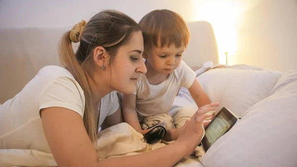 Retrato de madre joven con hijo pequeño acostado en la cama y usando tableta digital —  Fotos de Stock