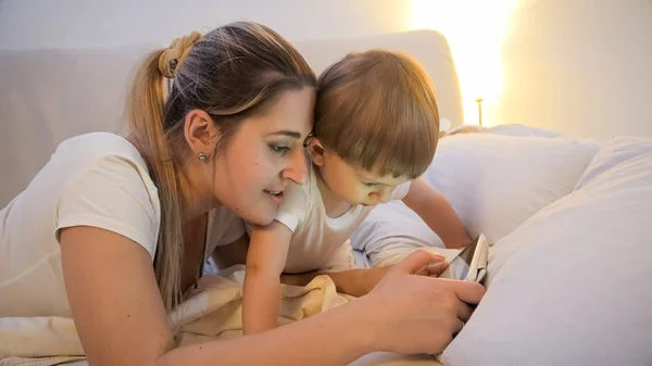 Portrait of young mother with her toddler son watching cartoons on digital tablet in bed