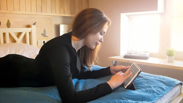 Hermosa joven mujer de negocios acostada en la cama en la habitación del hotel y utilizando la tableta digital —  Fotos de Stock