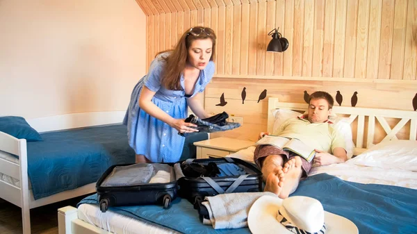 Ung kvinna packning saker för sommarsemester medan hennes make är liggande på sängen — Stockfoto