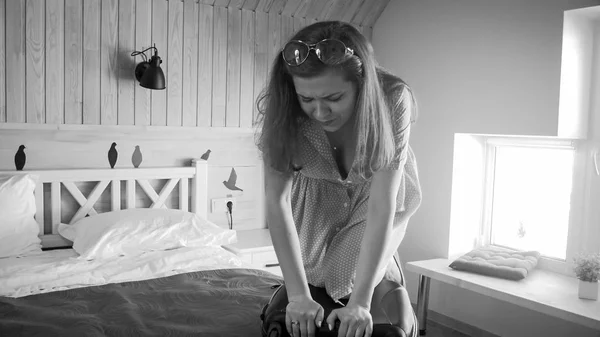 Black and white image of young woman sitting on suitcase trying to close it — Stock Photo, Image