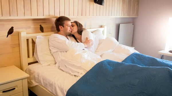 Retrato de jovem mulher bonita beijando seu marido deitado na cama no quarto de hotel — Fotografia de Stock