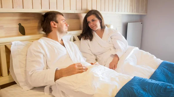 Happy young couple in bathrobes lying in bed and looking at each other — Stock Photo, Image