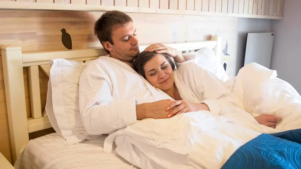 Portret van de jonge mens in badjas in bed liggen en zijn vrouw knuffelen — Stockfoto