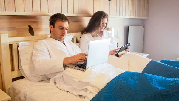 Mladý pár leží v posteli a pracuje na notebooku a tabletu počítače — Stock fotografie