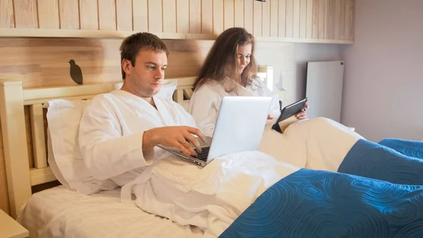 Jonge busy paar liggend in bed in de ochtend en het gebruik van de laptop en tablet pc — Stockfoto