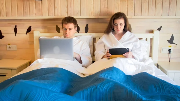Portrét mladý velmi rušné pár leží v posteli a pracuje na notebooku a tabletu počítače — Stock fotografie