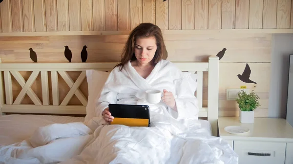 Retrato de una joven morena acostada en la cama y usando una tableta digital —  Fotos de Stock