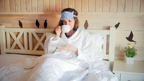 Portrét mladé ženy v županu seděl v posteli a pít čaj — Stock fotografie