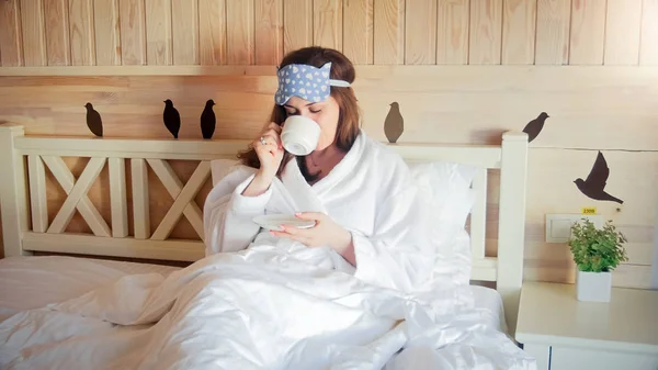 Portrét mladé ženy v županu s kávou v posteli ráno — Stock fotografie