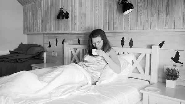 Imagen en blanco y negro de una joven tomando café en la cama por la mañana — Foto de Stock