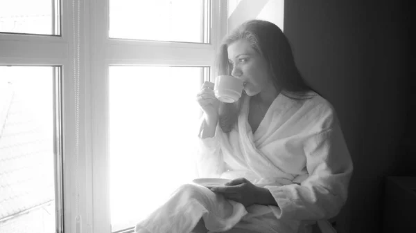 Portrait noir et blanc de jeune femme assise à la fenêtre et buvant du thé — Photo