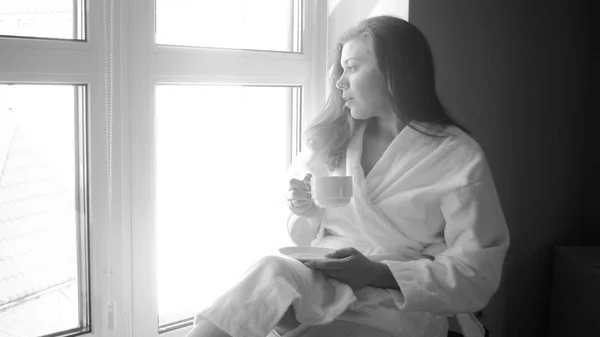 Schwarz-Weiß-Foto einer jungen Frau im Bademantel, die am großen Fenster Kaffee trinkt — Stockfoto