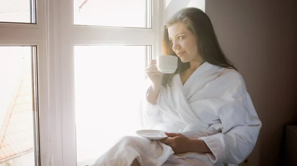 Retrato de una joven sonriente sentada en el alféizar de la ventana y bebiendo té — Foto de Stock
