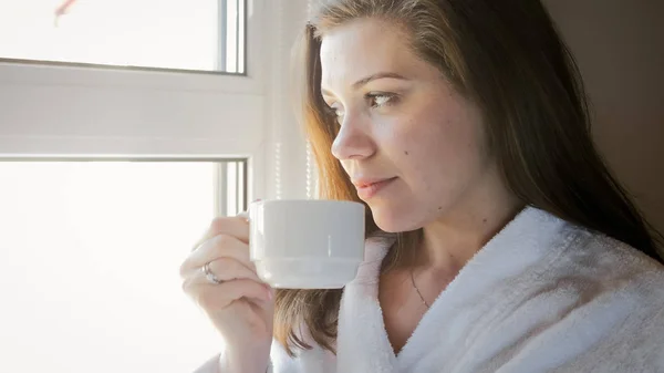 Retrato de una joven sonriente en albornoz mirando por la ventana y bebiendo té — Foto de Stock