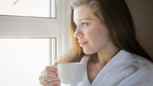 Potret wanita muda yang tersenyum duduk di jendela dan minum kopi — Stok Foto