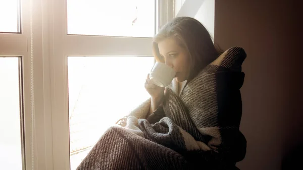 Hermosa mujer joven cubierta de cuadros sentada en la ventana bebiendo té — Foto de Stock