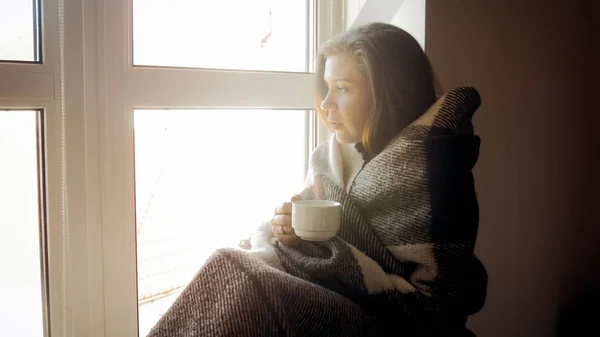 Young woman in plaid sitting on windowsill and drinking tea — Stock Photo, Image