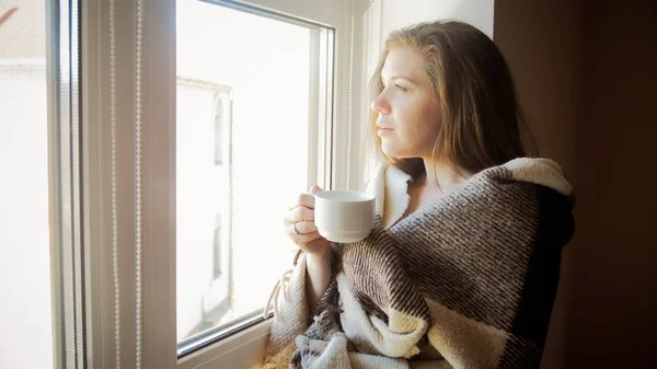 Крупный план тонированного портрета улыбающейся молодой женщины, покрытой клеткой с чашкой чая, стоящей у окна — стоковое фото