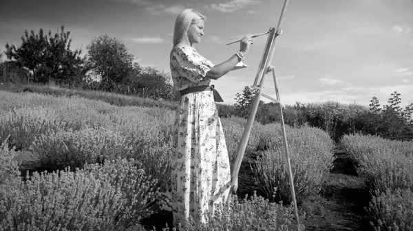 Black and white image of young woman painting landscape in field — Stock Photo, Image