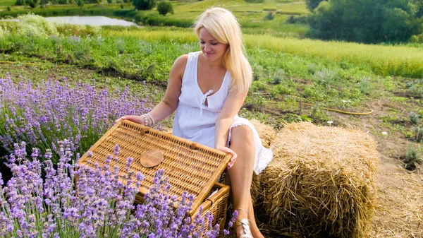 Mujer sonriente beautful sentado en la pila de heno y la apertura de la cesta de mimbre grande para el picnic — Foto de Stock