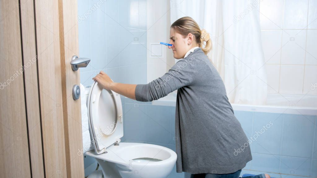 Young woman with clothespin on nose cleaning toilet at home