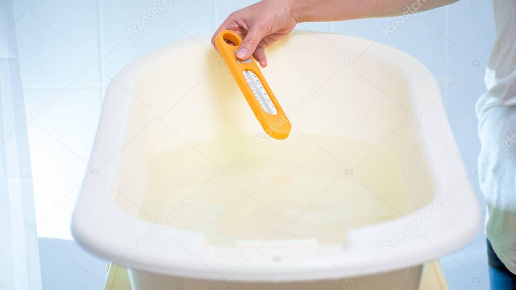 Closeup image of young woman checking water temperature with thermometer in baby bathtub