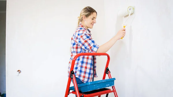 Portret van lachende jonge vrouw in hemd schilderen muren met verfroller — Stockfoto