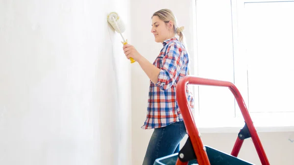 Mujer joven con camisa a cuadros y jeans pintando paredes en casa nueva —  Fotos de Stock