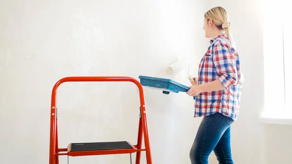 Hermosa mujer rubia joven woth rodillo de pintura y cubeta de pintura haciendo renovación en casa — Foto de Stock