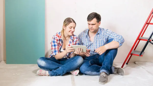 Щаслива молода пара сидить на підлозі в будинку під будівництвом і використовує цифровий планшет — стокове фото