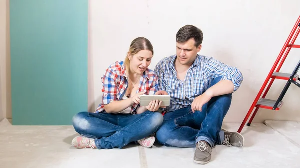 Glada unga paret sitter på golvet vid hus under renovering och surfar internet på digitala tablett — Stockfoto
